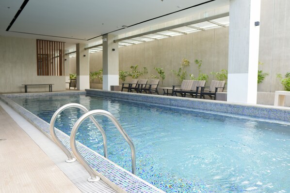 An indoor swimming pool with clear water, metal pool ladders, and several lounge chairs around the pool area.