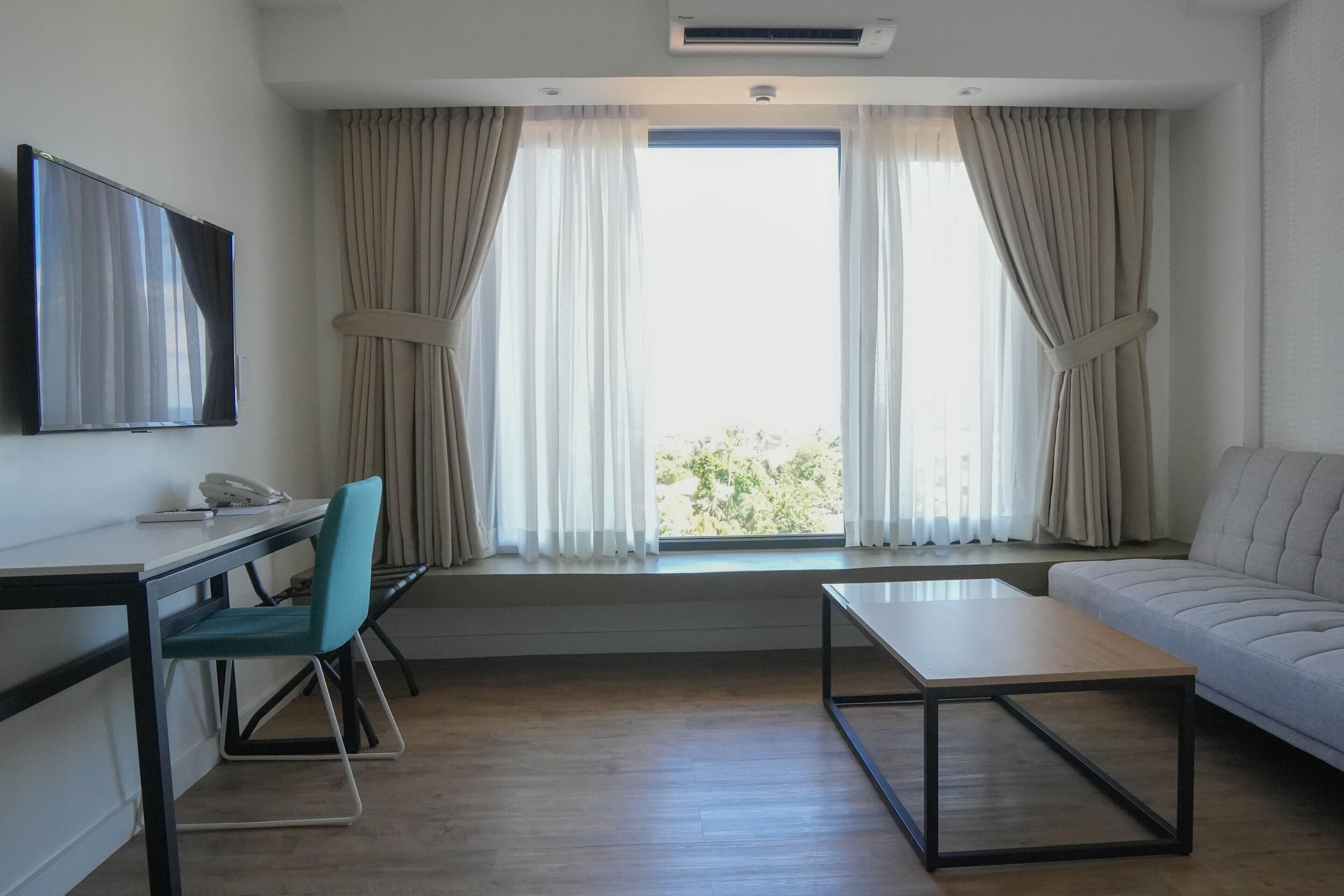 A modern hotel room with a desk, chair, sofa, and a large window with curtains.