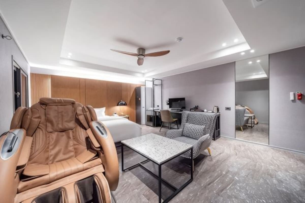 A modern hotel room with a massage chair, a bed, a desk, and a seating area.