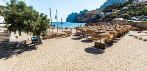 Een strand met rieten parasols, een heldere blauwe zee en een rotsachtige klif op de achtergrond.