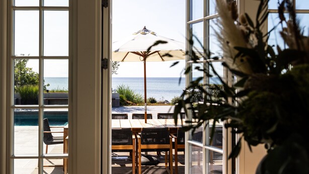 An open door leading to a patio with a table, chairs, and an umbrella overlooking a pool and the sea. - Oakvale Villa Sand (Aahus)