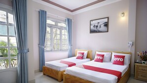 A hotel room with two beds, a window with curtains, a bedside table with a lamp, and a painting on the wall.