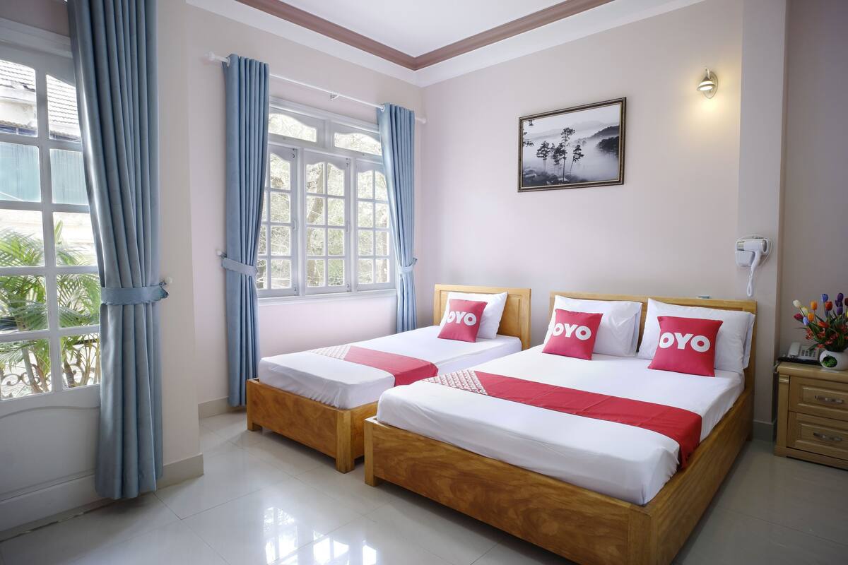 Une chambre d’hôtel avec deux lits, une fenêtre avec des rideaux, une table de chevet avec une lampe et un tableau au mur.