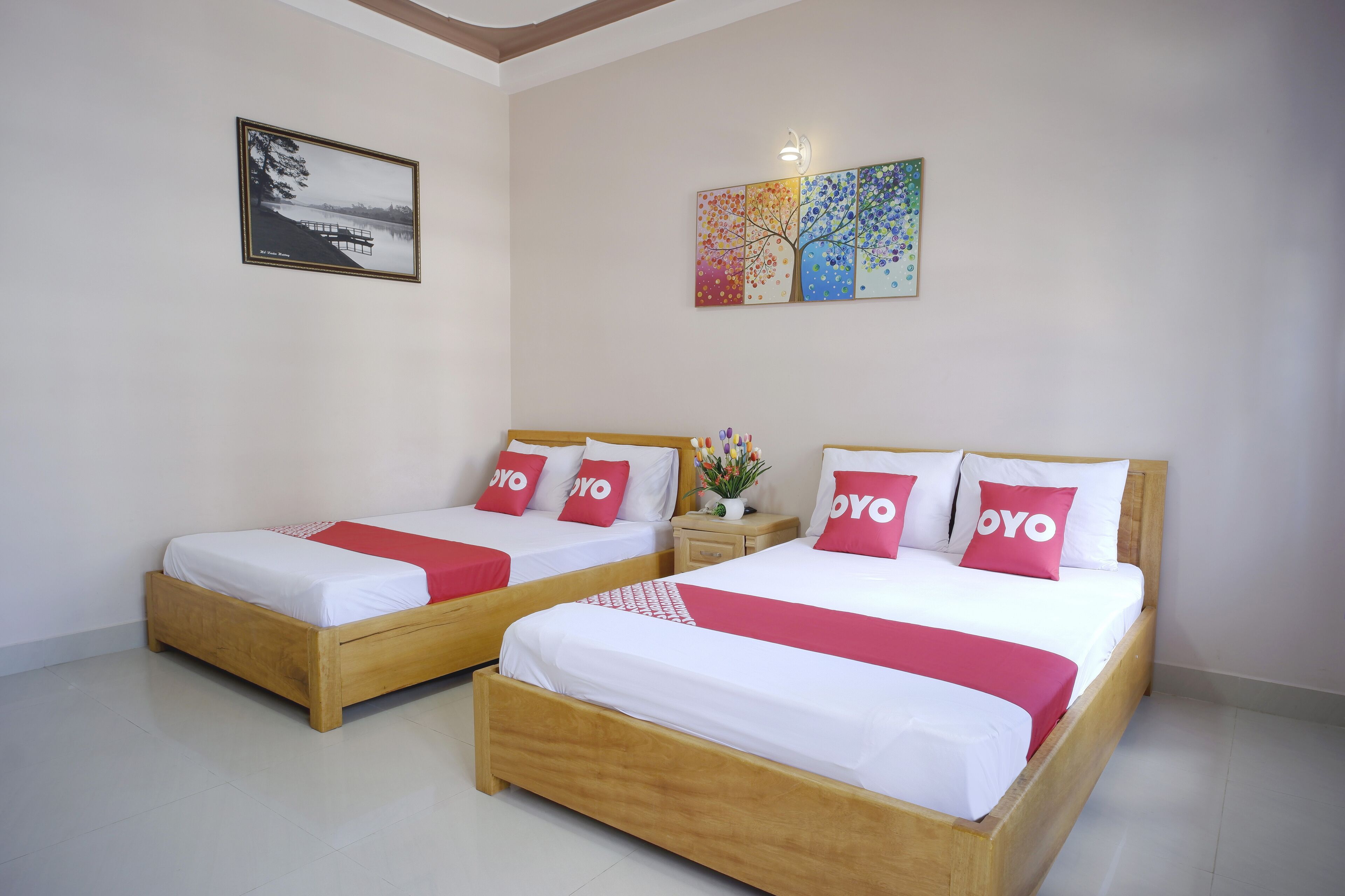 Ein Hotelzimmer mit zwei hölzernen Betten, jeweils mit weißer Bettwäsche und roten Akzenten, darunter Kissen und eine Decke. An der Wand hängt ein gerahmtes Bild und über den Betten ein farbenfrohes Kunstwerk.