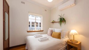A neatly made bed with folded towels, a bedside lamp, a window with a view of buildings, and a shelf with a plant.