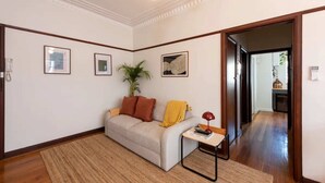 A living room with a sofa, a small table, and a plant. There are framed pictures on the wall and a door leading to another room.