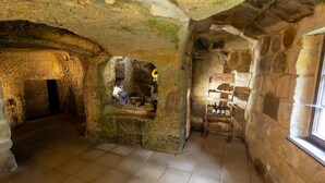 Ein Zimmer mit Steinwänden, gefliestem Boden, einem Holzregal mit Flaschen und einem kleinen Fenster.