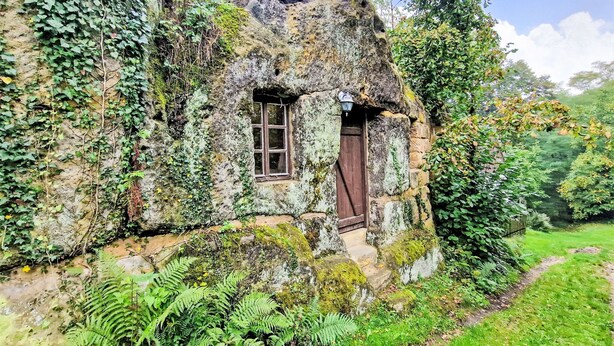 Ein Steinhäuschen mit kleinem Fenster und Tür, umgeben von üppigem Grün und Farne.