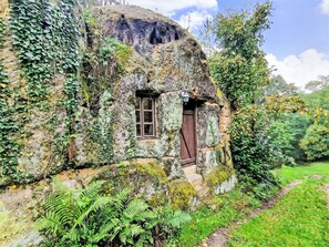 A stone cottage with a small window and door, surrounded by lush greenery and ferns.