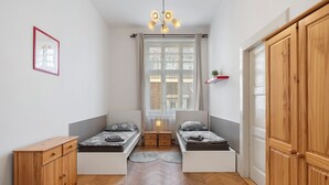A compact bedroom with two single beds, a wooden wardrobe, a small desk, and a window with a view of buildings.