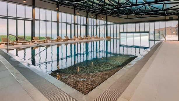 An indoor swimming pool with clear glass walls, lounge chairs, and a view of the landscape.