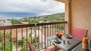 Un balcon avec une table dressée pour deux personnes, une chaise et une bouteille de jus. La vue offre un paysage côtier avec des maisons et de la verdure.