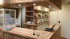 Un mostrador de recepción con una computadora portátil, un teléfono y un dispensador de agua. Hay estantes con plantas en macetas y un cartel que indica los servicios.