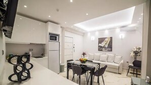 Una cocina moderna con armarios blancos, un área de comedor con mesa y sillas negras, y un sofá blanco en un espacio luminoso con tragaluces.
