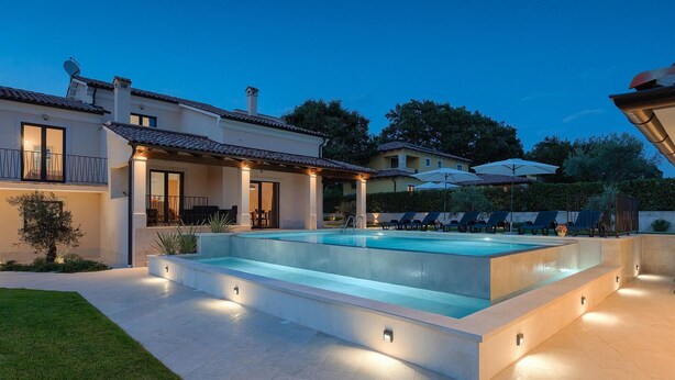 A modern outdoor pool area with a rectangular pool, surrounded by a light-colored deck, and a covered patio with outdoor furniture.