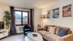 A living room with a sofa, a coffee table, a chair, a TV, a radiator, a window with curtains, and a plant.