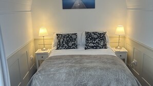 Una cama bien hecha con una colcha gris, dos lámparas de noche y un cuadro de un muelle en la pared.