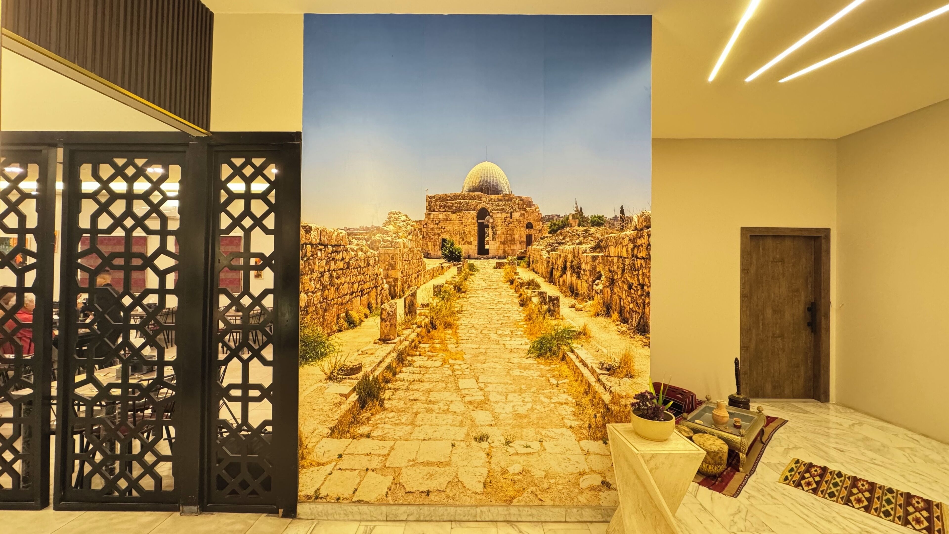 Una habitación con un amplio mural histórico en la pared, una puerta de madera y una mesita de noche de mármol con un jarrón y un cuenco.