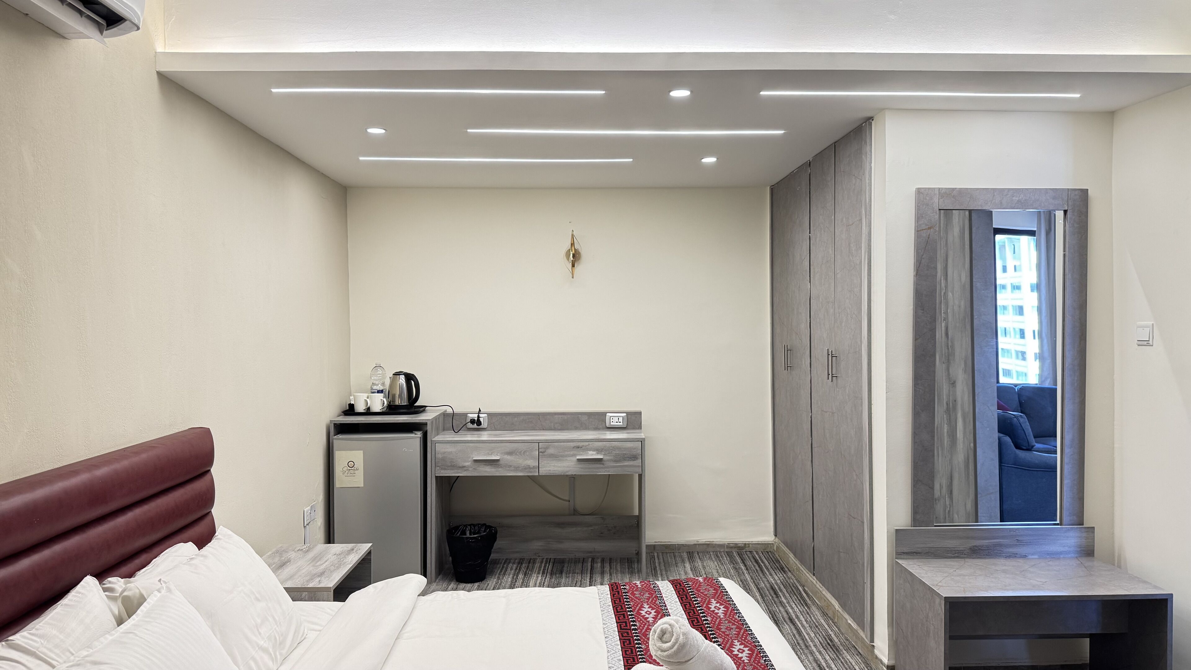 A hotel room with a bed, a desk with a kettle, a small fridge, a wardrobe, and a view of the outside.