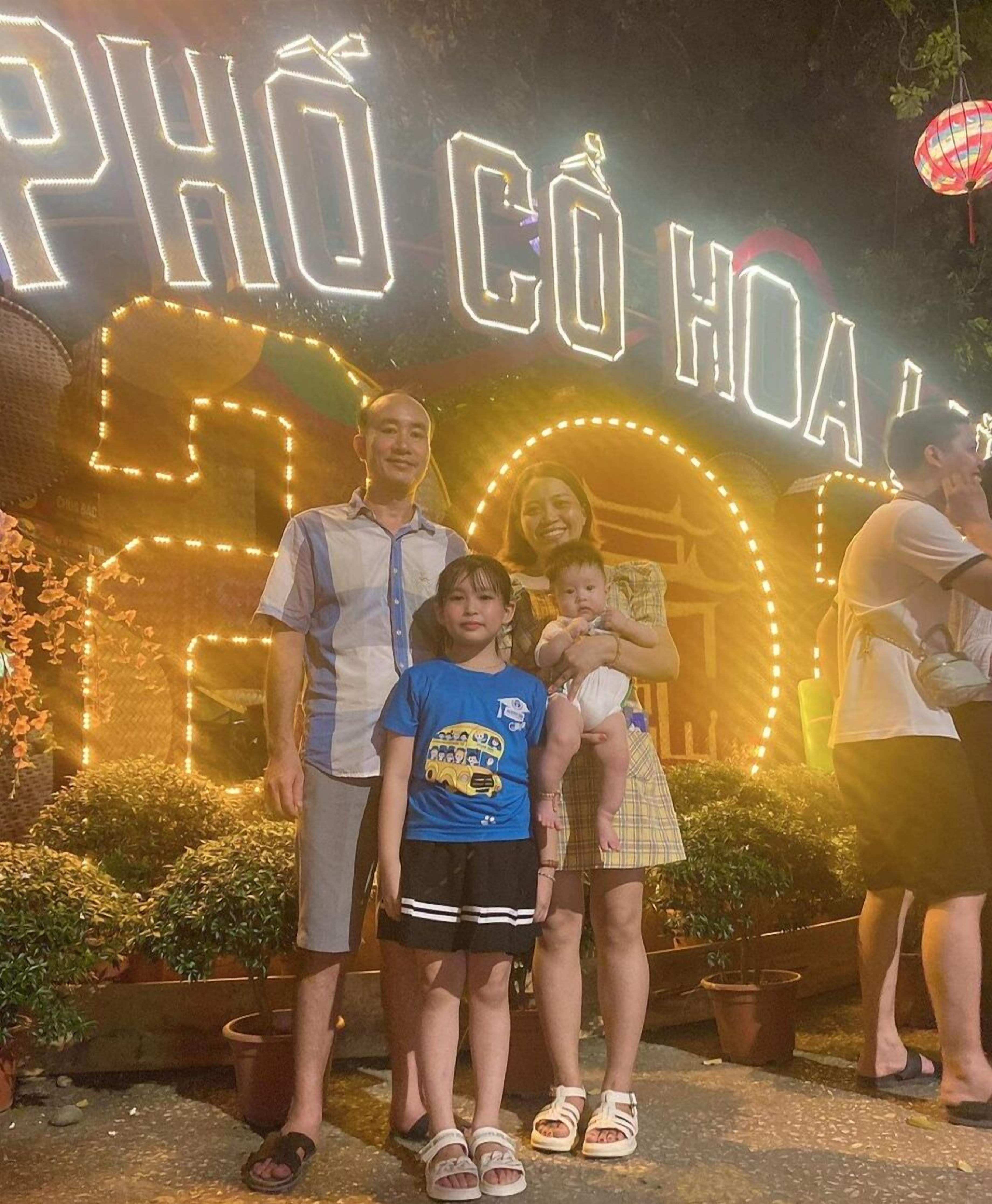 Une famille pose devant une enseigne au néon sur laquelle on peut lire le texte « PHÒ CÔ HỒI » et le chiffre « 50 ».