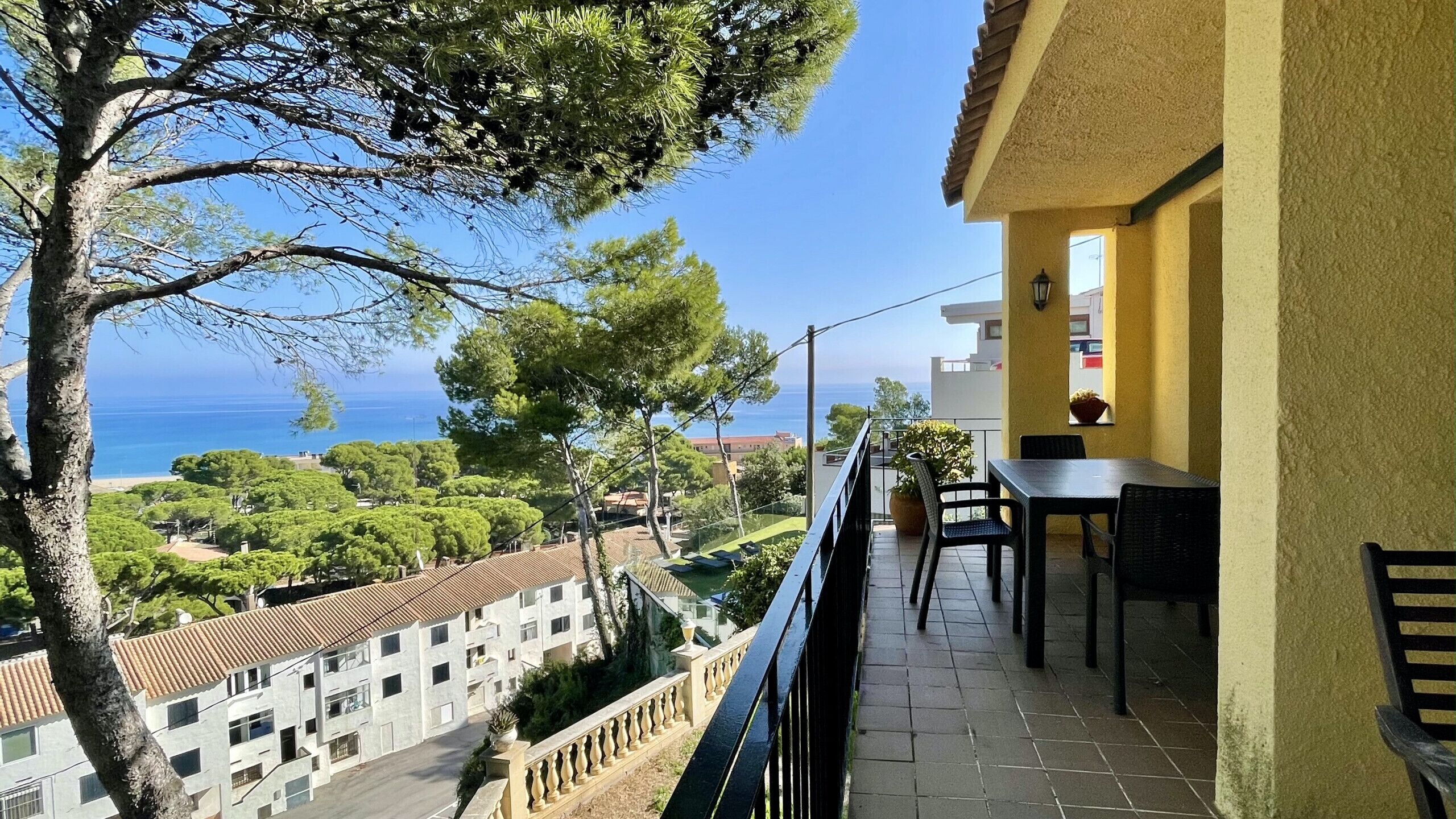 A balcony with a table and chairs, overlooking a coastal area with buildings and trees.