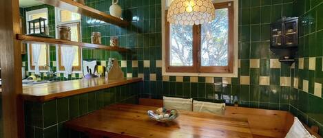 Una cocina con baldosas verdes, una mesa de madera, una lámpara colgante y una ventana con vistas a árboles.