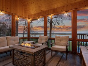 Una terraza de madera con chimenea de piedra, asientos acolchados y grandes ventanales con vista a un lago al atardecer.