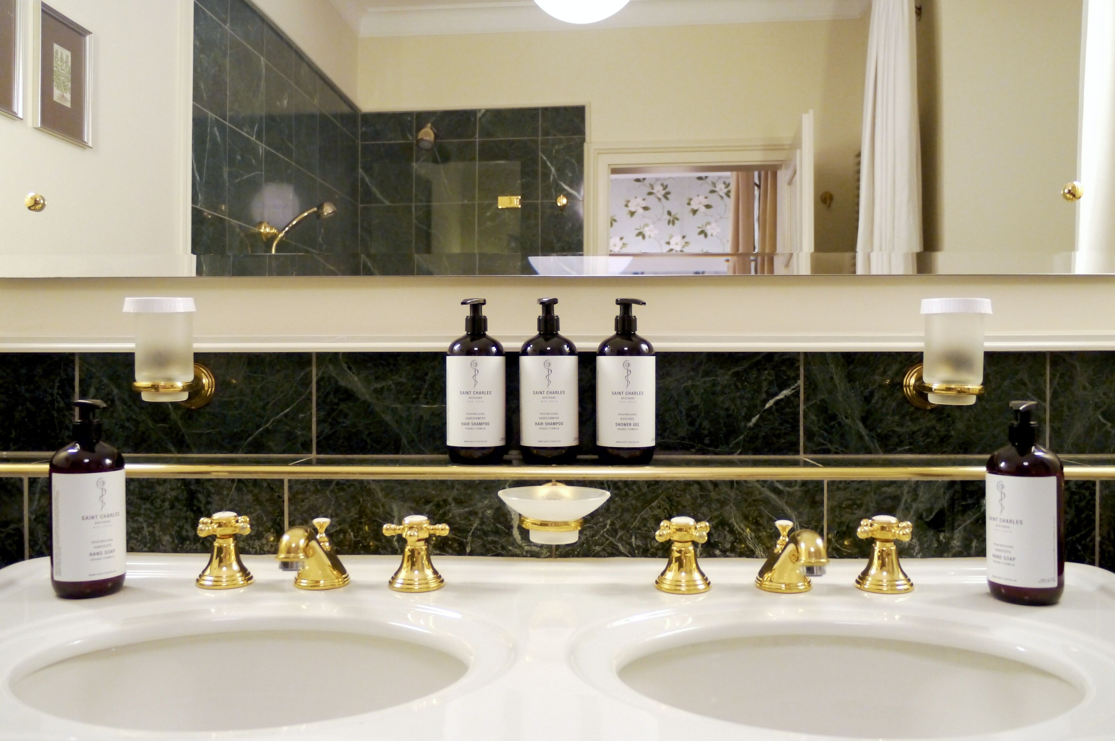 Un baño con dos lavamanos, grifos dorados y tres botellas de gel de ducha Saint Charles sobre el mostrador.