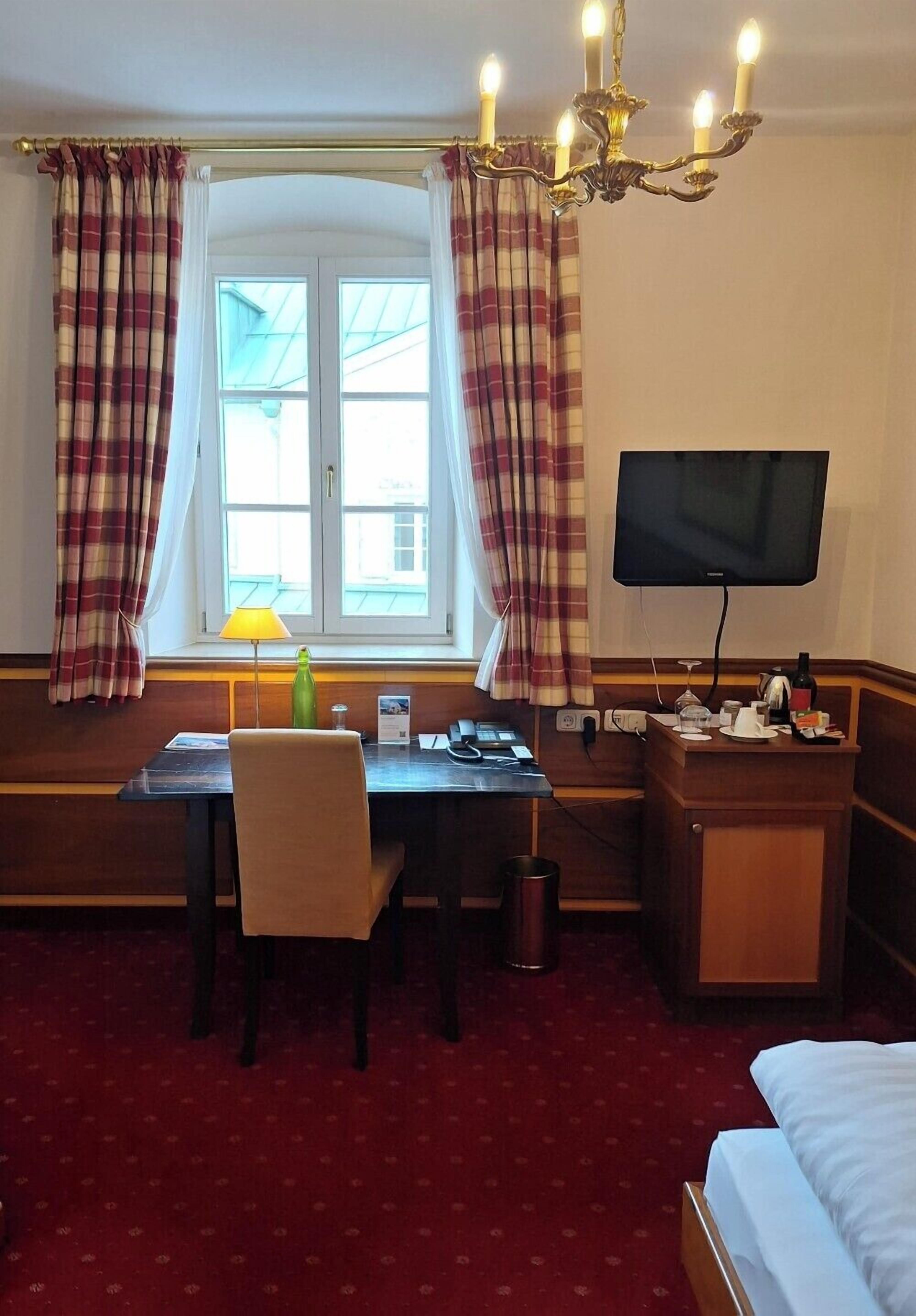 Habitación de hotel con cama, escritorio, silla, televisor, lámpara y ventana con cortinas.