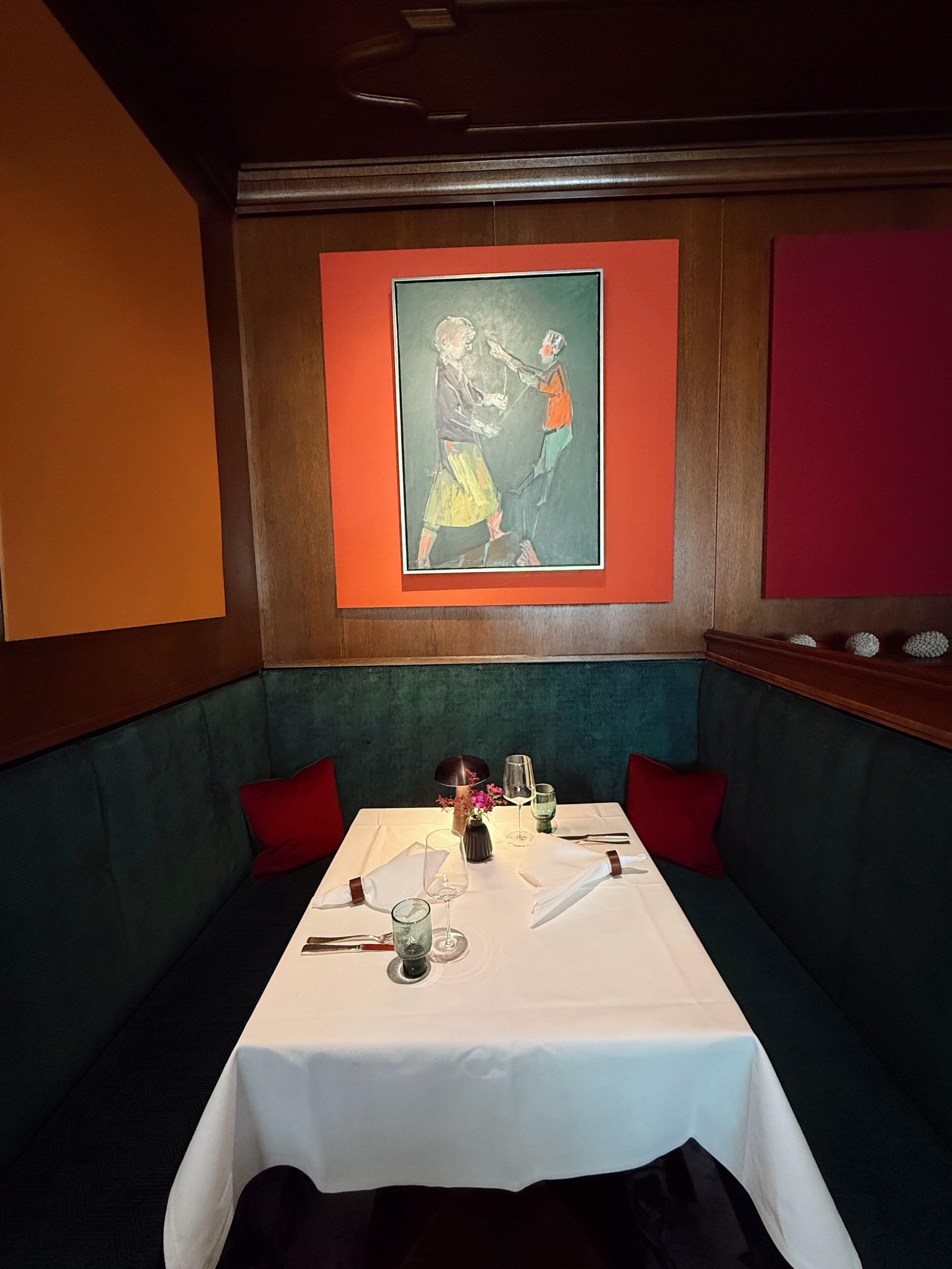 Uma área de refeições com uma mesa posta para dois, um quadro na parede e um banco estofado verde.
