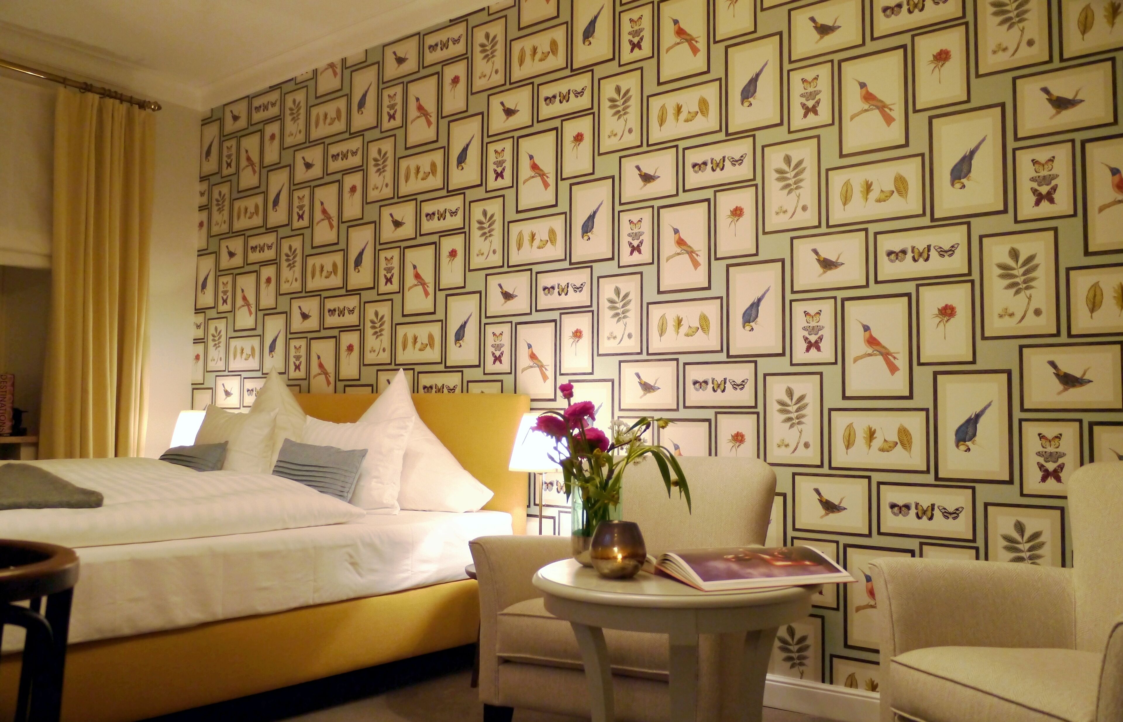 Una habitación de hotel con una cama, una silla, una mesita con un florero y una pared decorada con cuadros de pájaros y hojas.