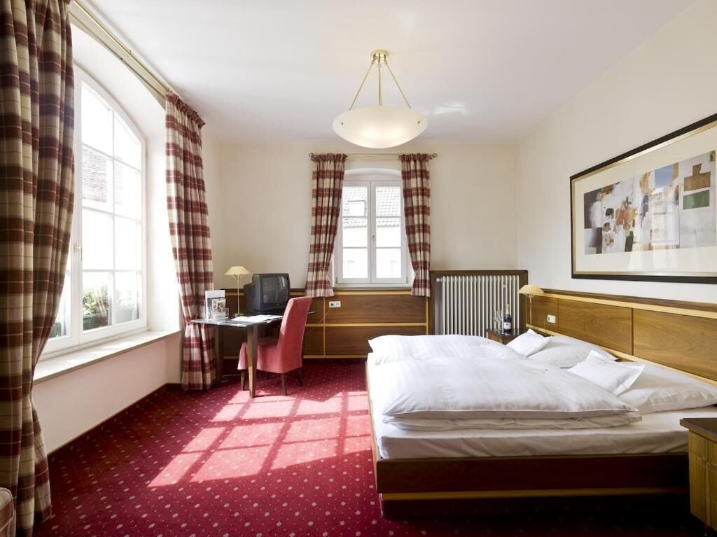 Et hotelværelse med en seng, et skrivebord med stol, en radiator og et vindue med gardiner.