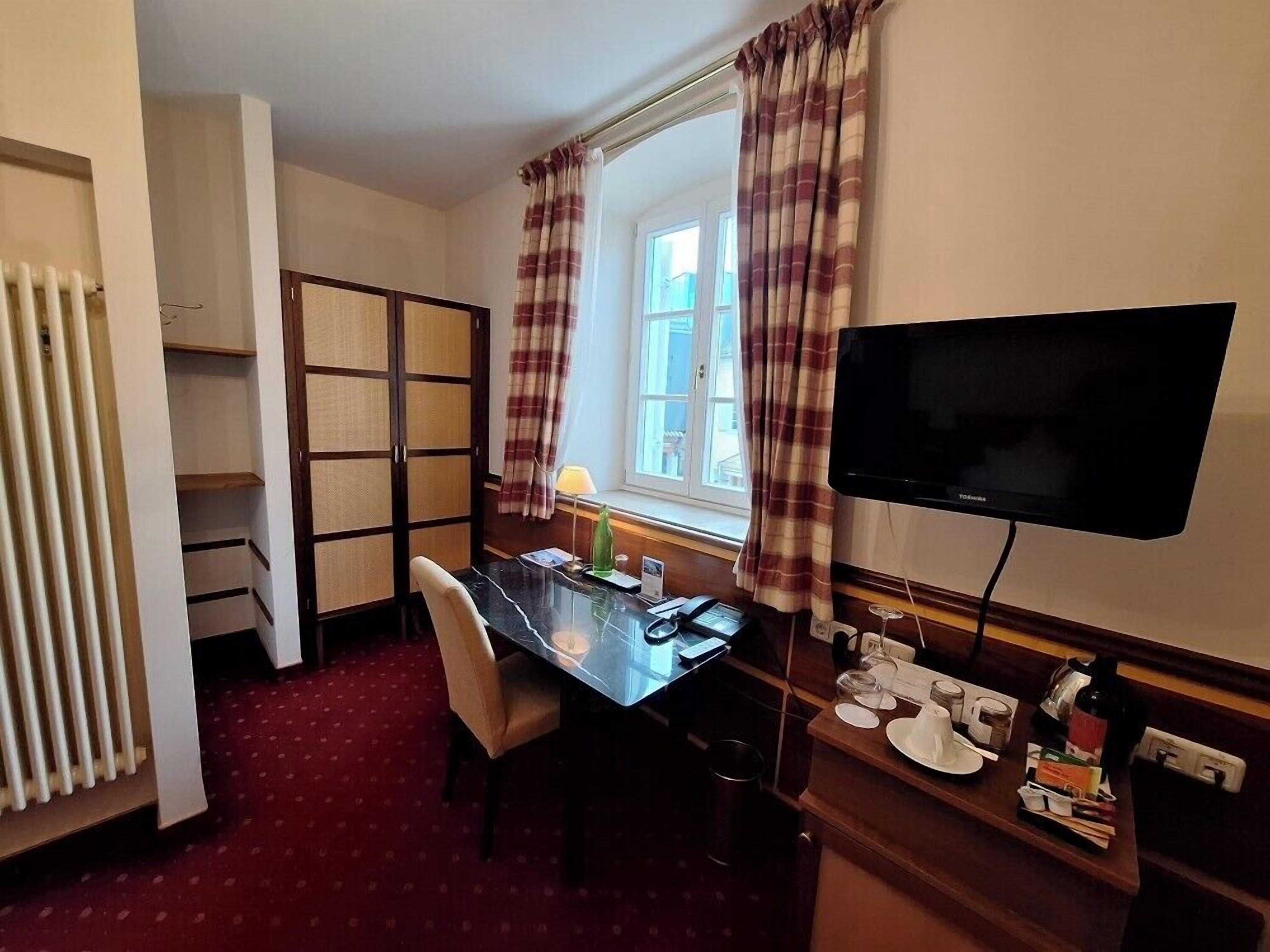 Una habitación de hotel con cama, escritorio con silla, televisor y ventana con cortinas.