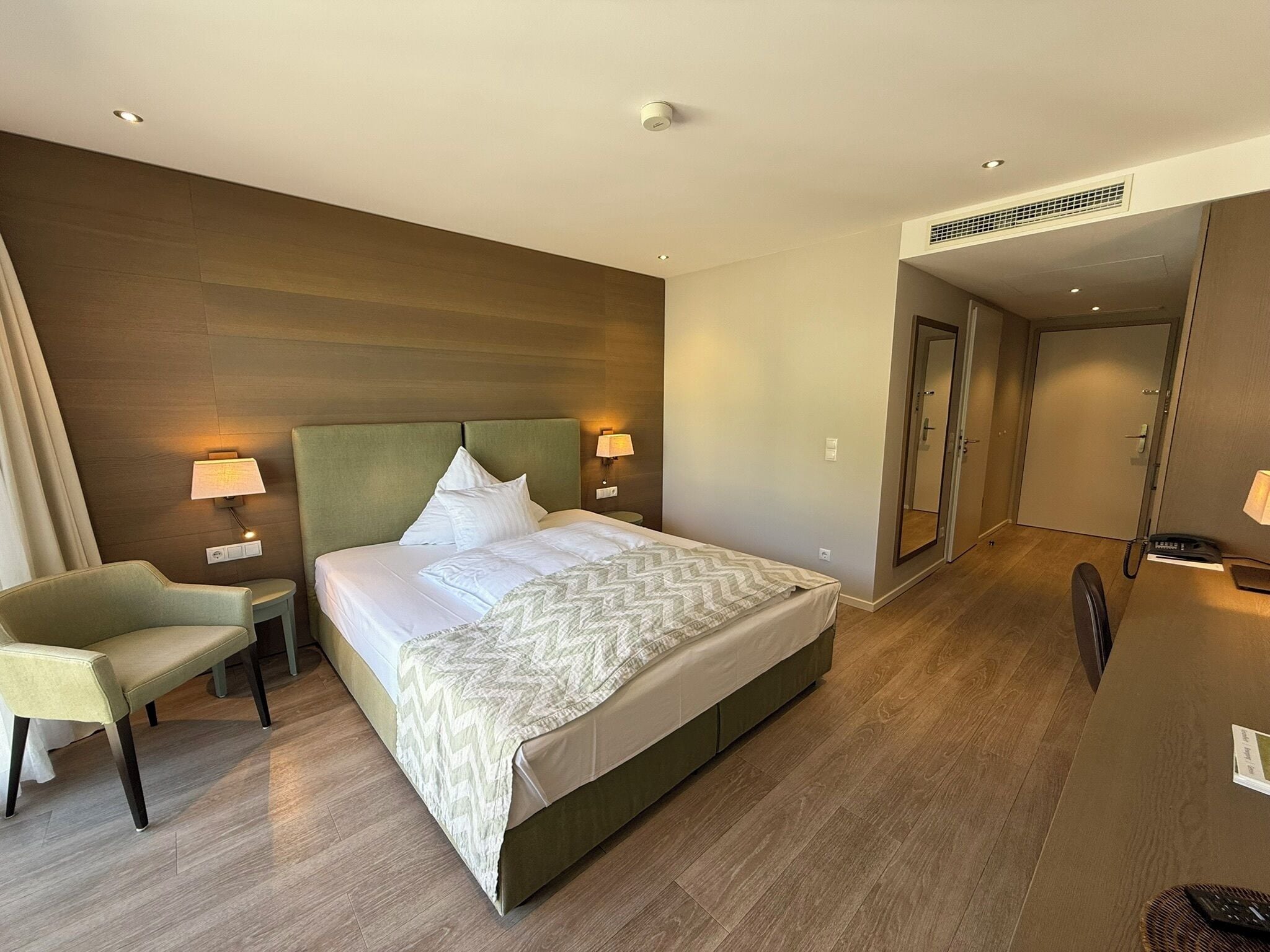 A hotel room with a large bed, two bedside lamps, a chair, a desk with a phone, and a door leading to another room.