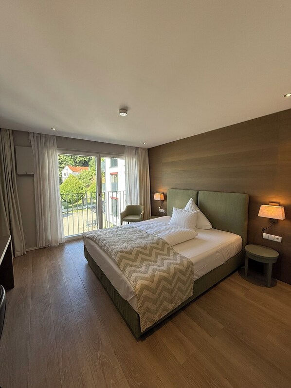 A hotel room with a large bed, a small bedside table, a chair, and a view of a balcony. - Romantik Hotel Das Lindner (Bad Aibling)