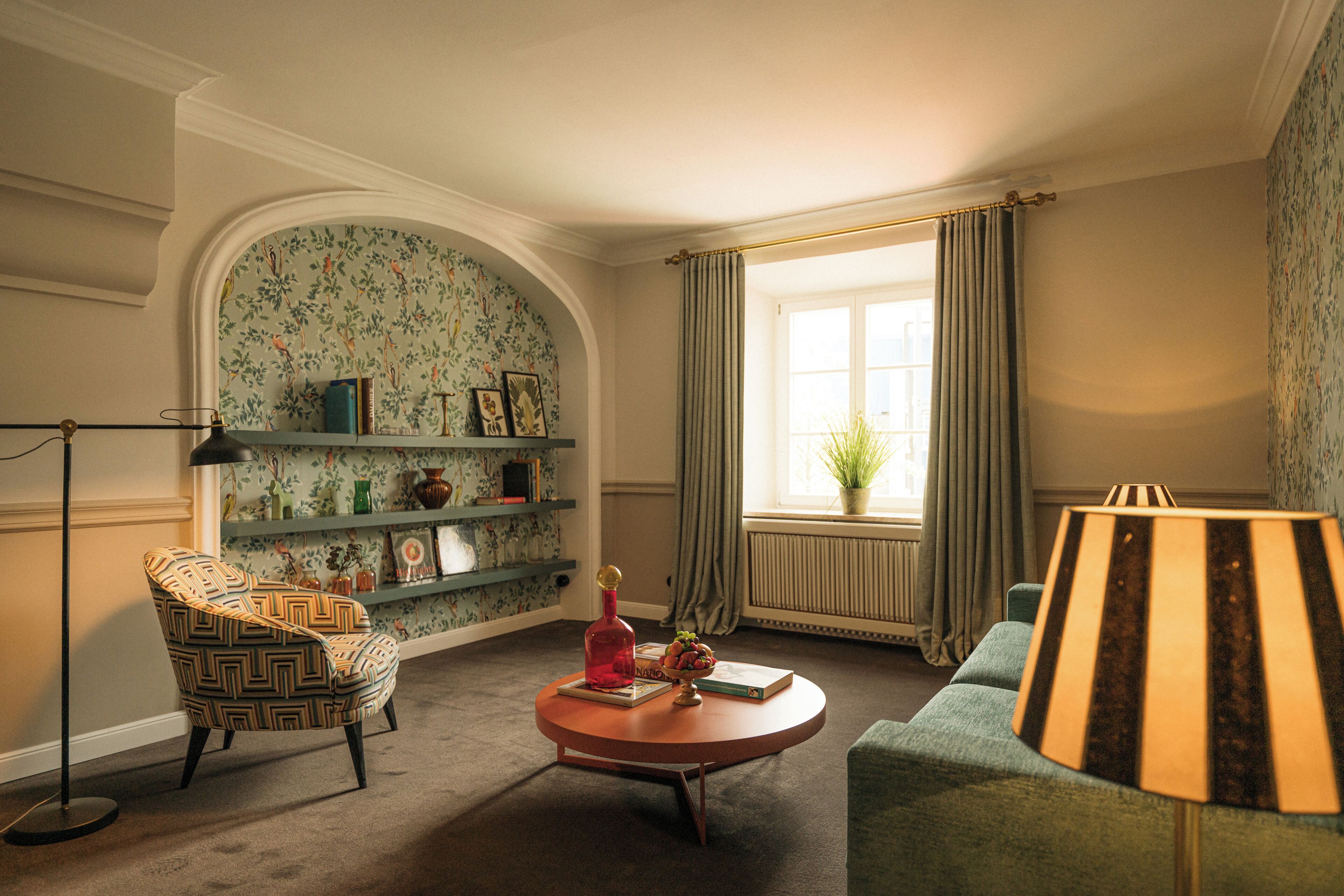 A room with a patterned wallpaper, a round coffee table, a patterned armchair, a sofa, a lamp, and a window with curtains.
