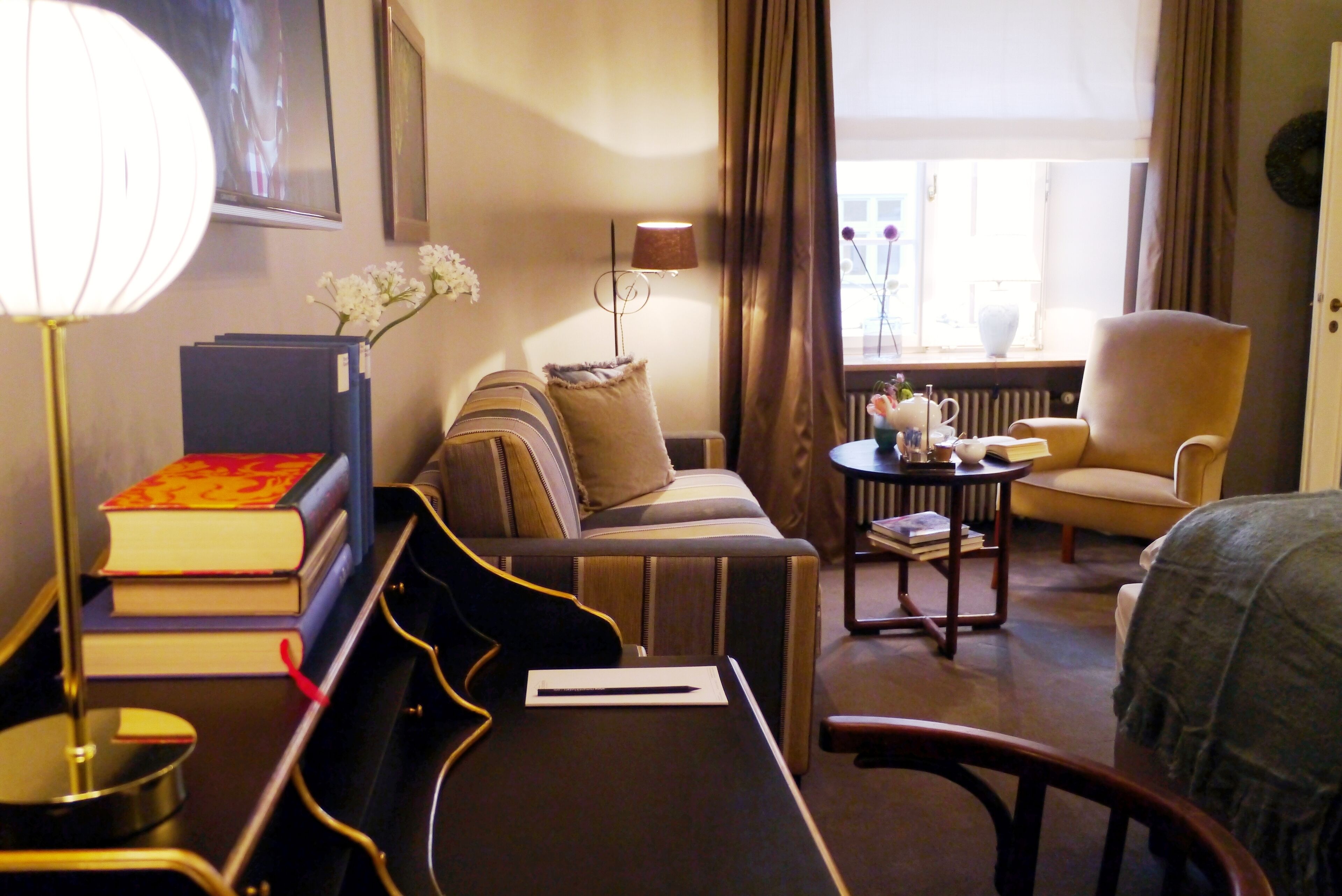A hotel room with a sofa, armchair, bedside table, lamp, and a window with curtains.