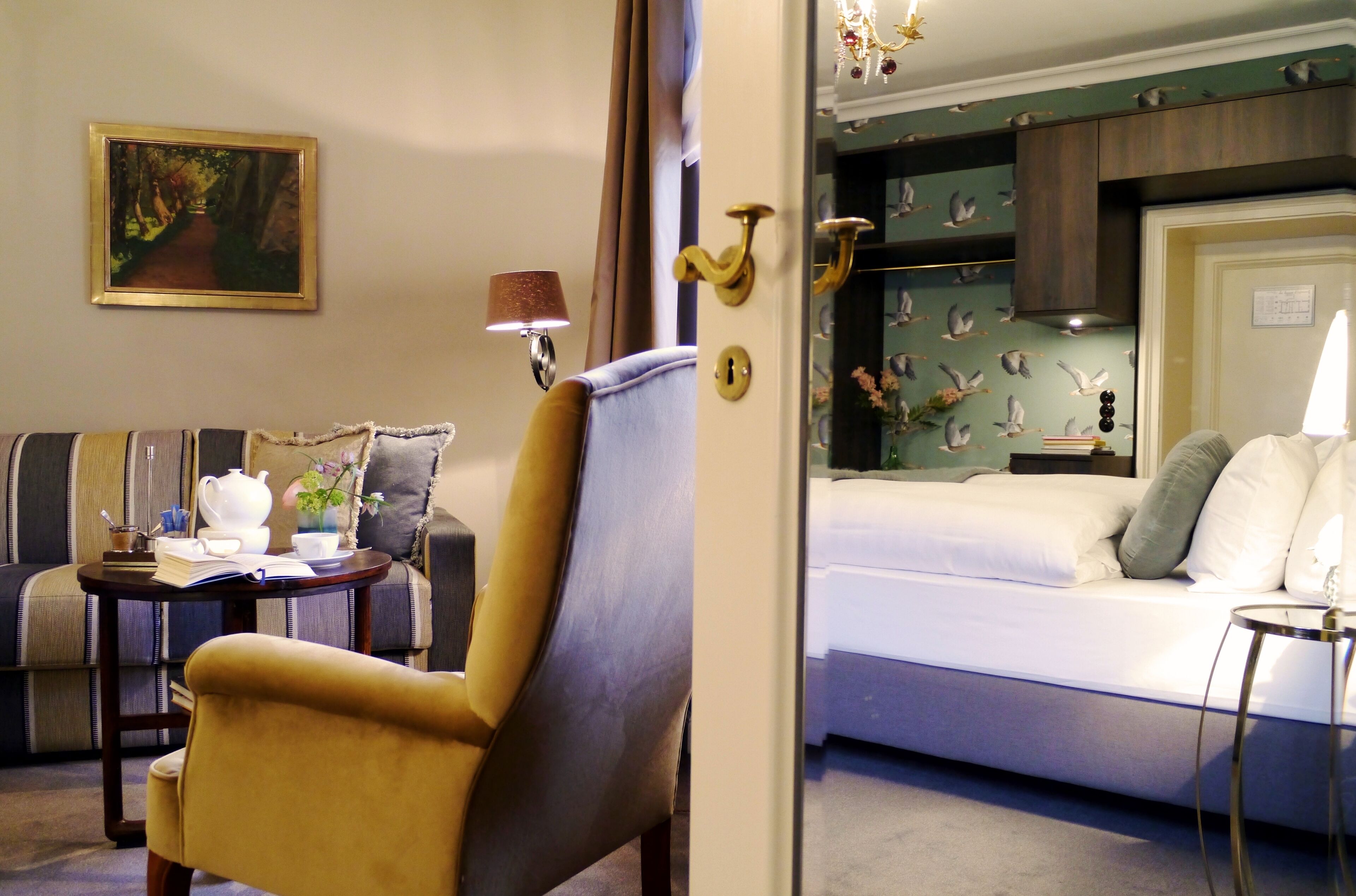 Habitación de hotel con sofá, sillón, mesita de noche, lámpara y cama con cabecero blanco.