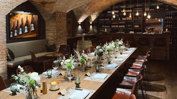 A long dining table with chairs, a brick wall, and a wine rack in the background. - Romantik Hotel Das Lindner (Bad Aibling)
