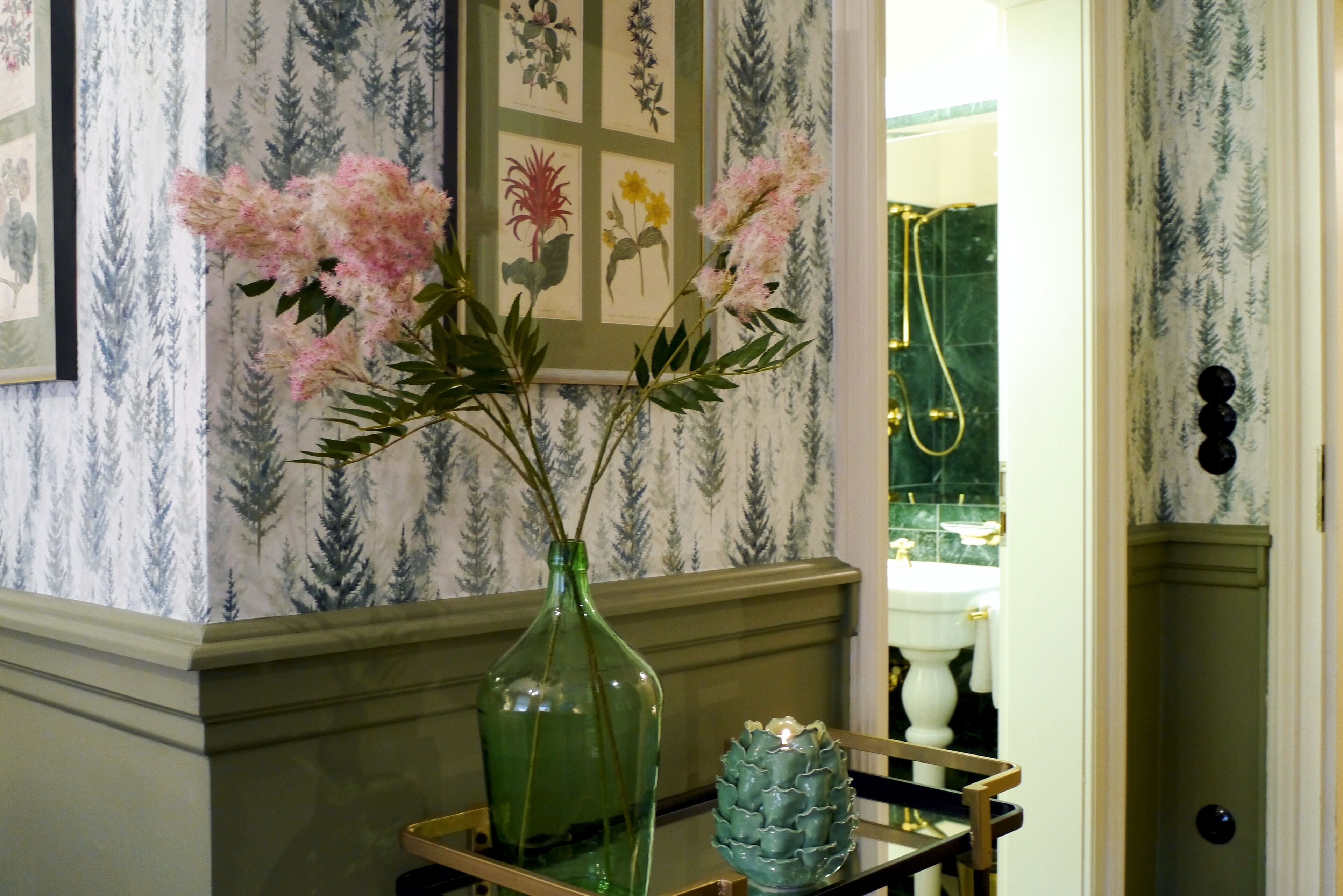 A bathroom with a green vase and pink flowers on a tray, wallpaper with a forest pattern, and a gold showerhead.