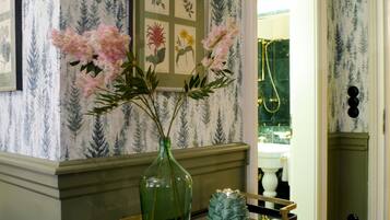 A bathroom with a green vase and pink flowers on a tray, wallpaper with a forest pattern, and a gold showerhead.
