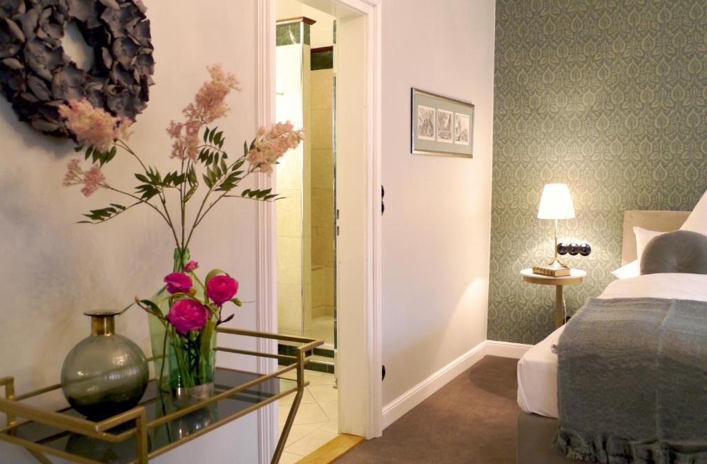 Una habitación de hotel con una cama, una mesita de noche con lámpara, un espejo, un arreglo de pared y un jarrón con flores.