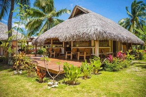 Una cabaña con techo de paja y terraza de madera, con zona de estar al aire libre, rodeada de follaje tropical.