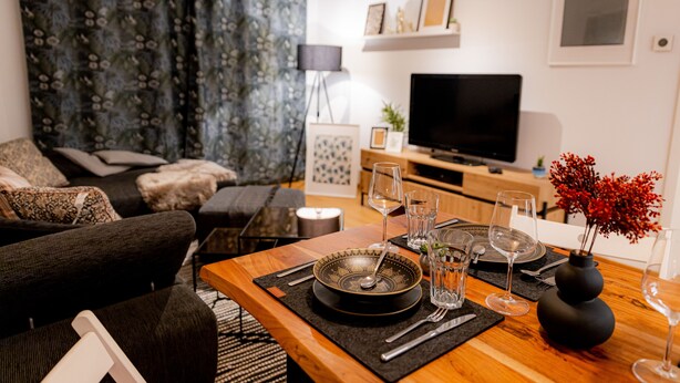 A modern living room with a sofa, a dining table set with plates, glasses, and cutlery, and a flat-screen TV mounted on the wall.