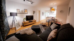 A modern living room with a sofa, ottoman, and a flat-screen TV on a wooden stand. There is a dining area with a table and chairs, and a kitchen in the background.