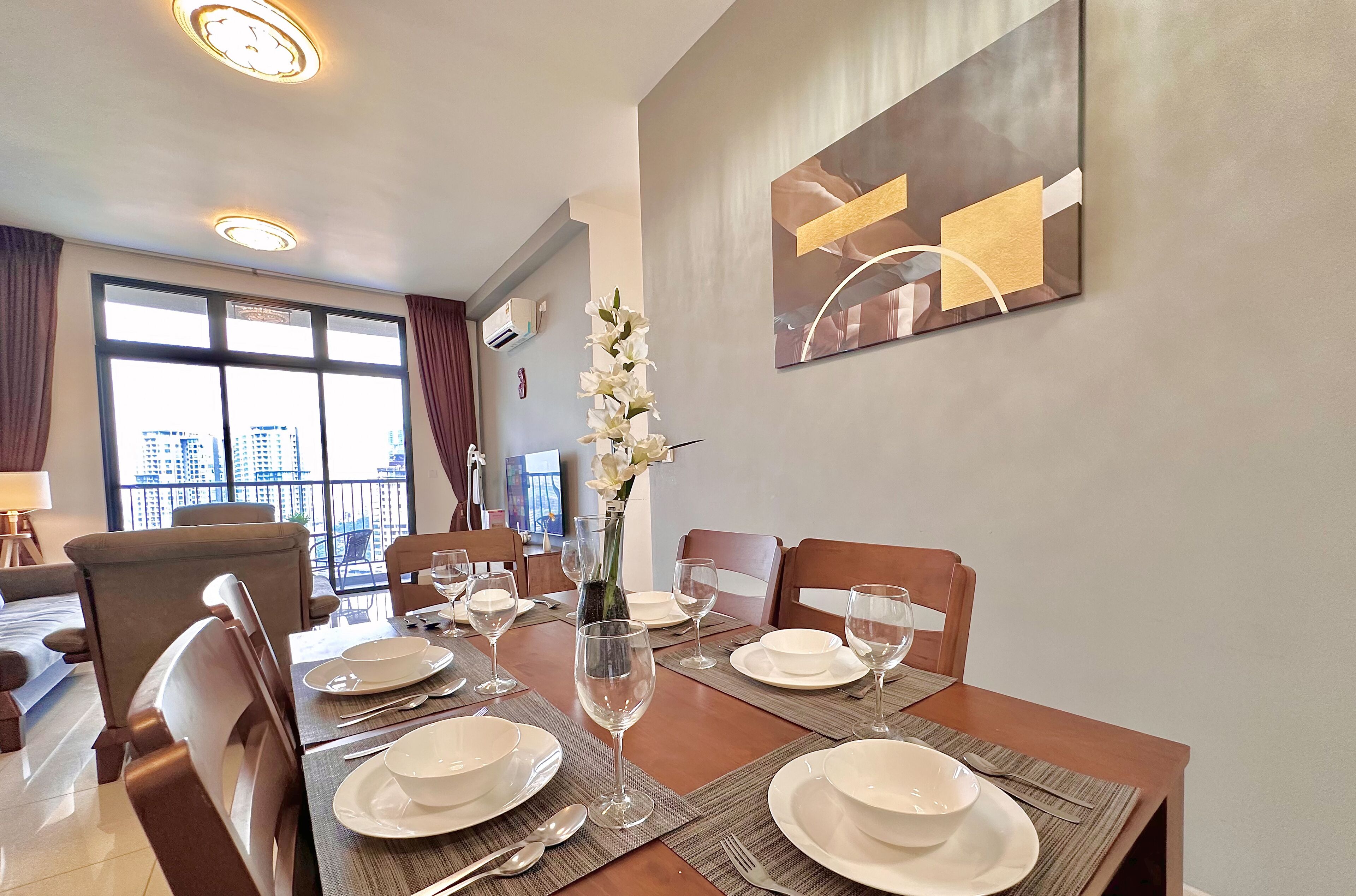 A dining area with a table set for four, a vase with white flowers, and a city view through large windows.