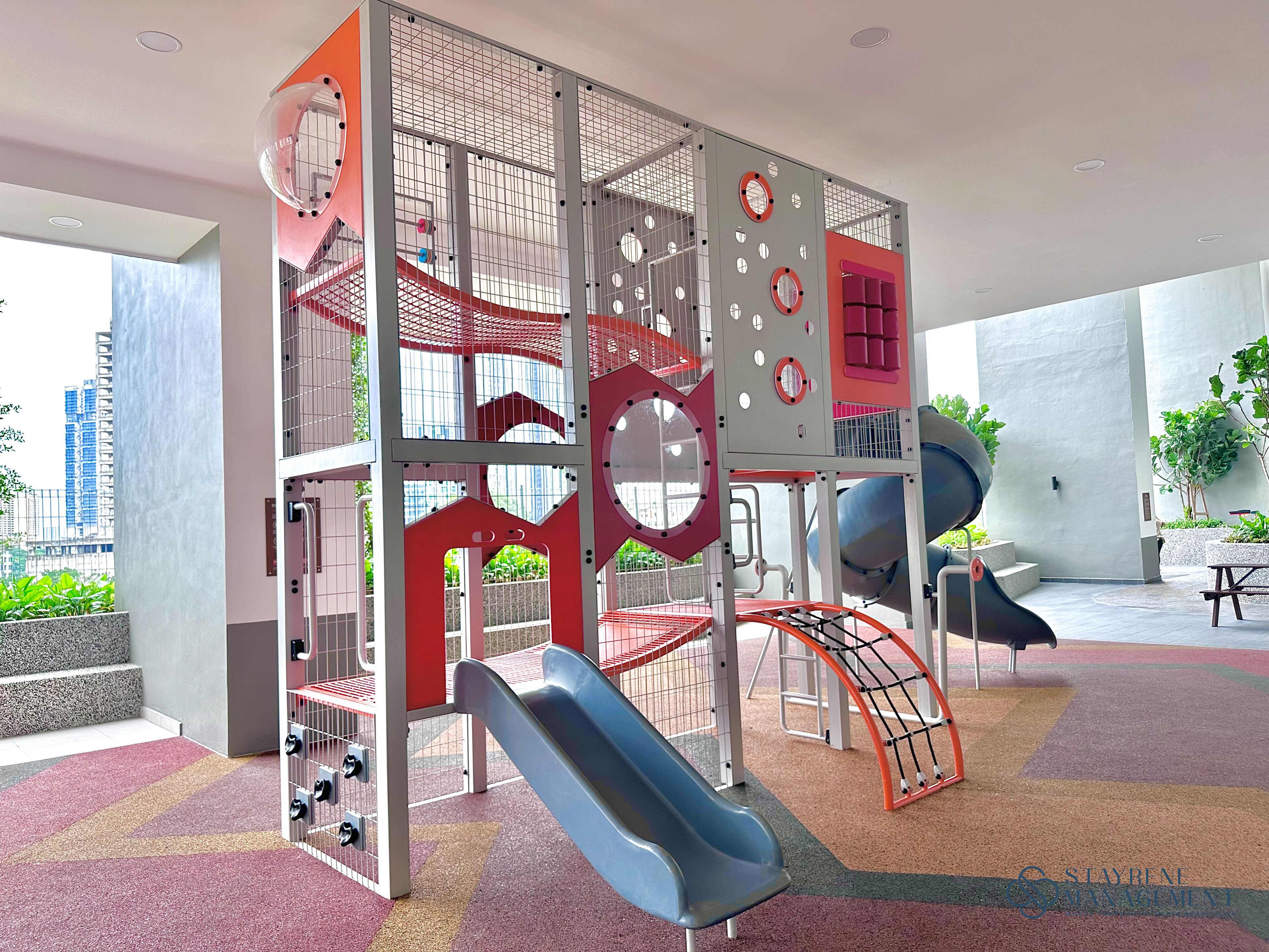 A multi-level playground with slides and climbing structures, surrounded by a modern building with large windows and a view of the cityscape.