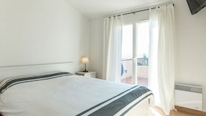 A hotel room with a bed, a nightstand, a lamp, a window with curtains, and a view of the outside. - Esprit du Sud Vakantiehuis met 3 Slaapkamers, Mooi Uitzicht op Domaine du Golf (Roquebrune-sur-Argens)