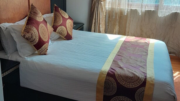 Una habitación de hotel con una cama grande, cabecera, mesita de noche, lámpara de pared y ventana con cortinas.