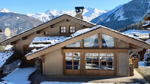 Eine Chalet mit geneigtem Dach, Holzbau und großen Fenstern. Umgeben ist es von schneebedeckten Bergen und wolkenlosem Himmel.